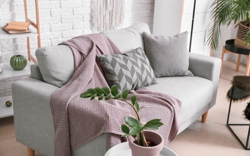 Canapé gris très clair, avec un plaid de couleur vieux rose, des coussins, on voit un mur en brique blanches derrière et une plante verte une une petite table sur le côté.