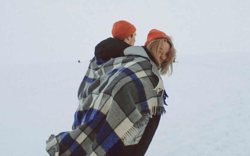 Deux personnes partageant un grand plaid à carreaux bleus et blancs, se réchauffant dans un paysage enneigé, portant des bonnets oranges assortis.