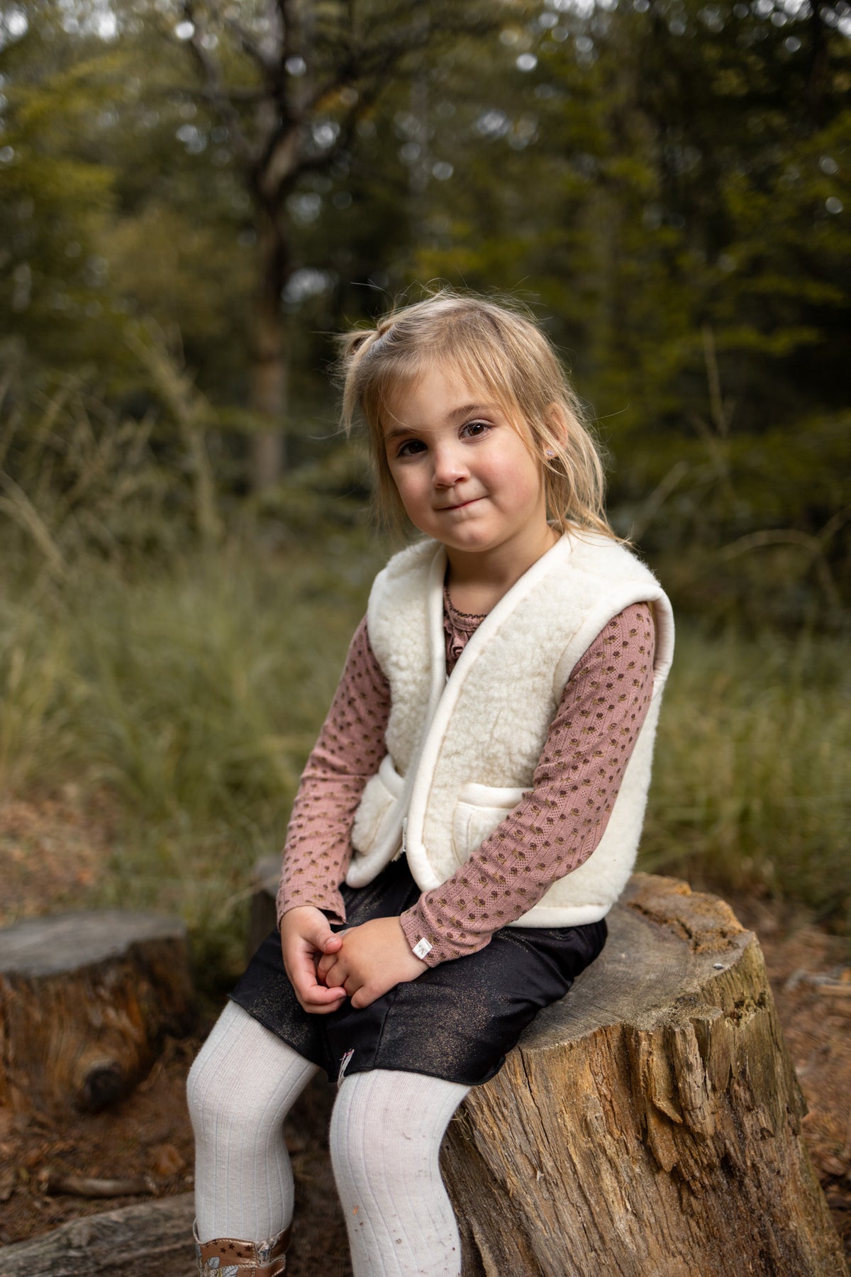 Gilet sans manches en laine pour enfant blanc cassé, porté par une petite fille assise sur une souche